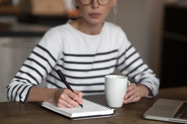 Elegant smart woman in glasses writing in spiral notepad journal or ideas placed on bright desktop with coffee cup in the morning